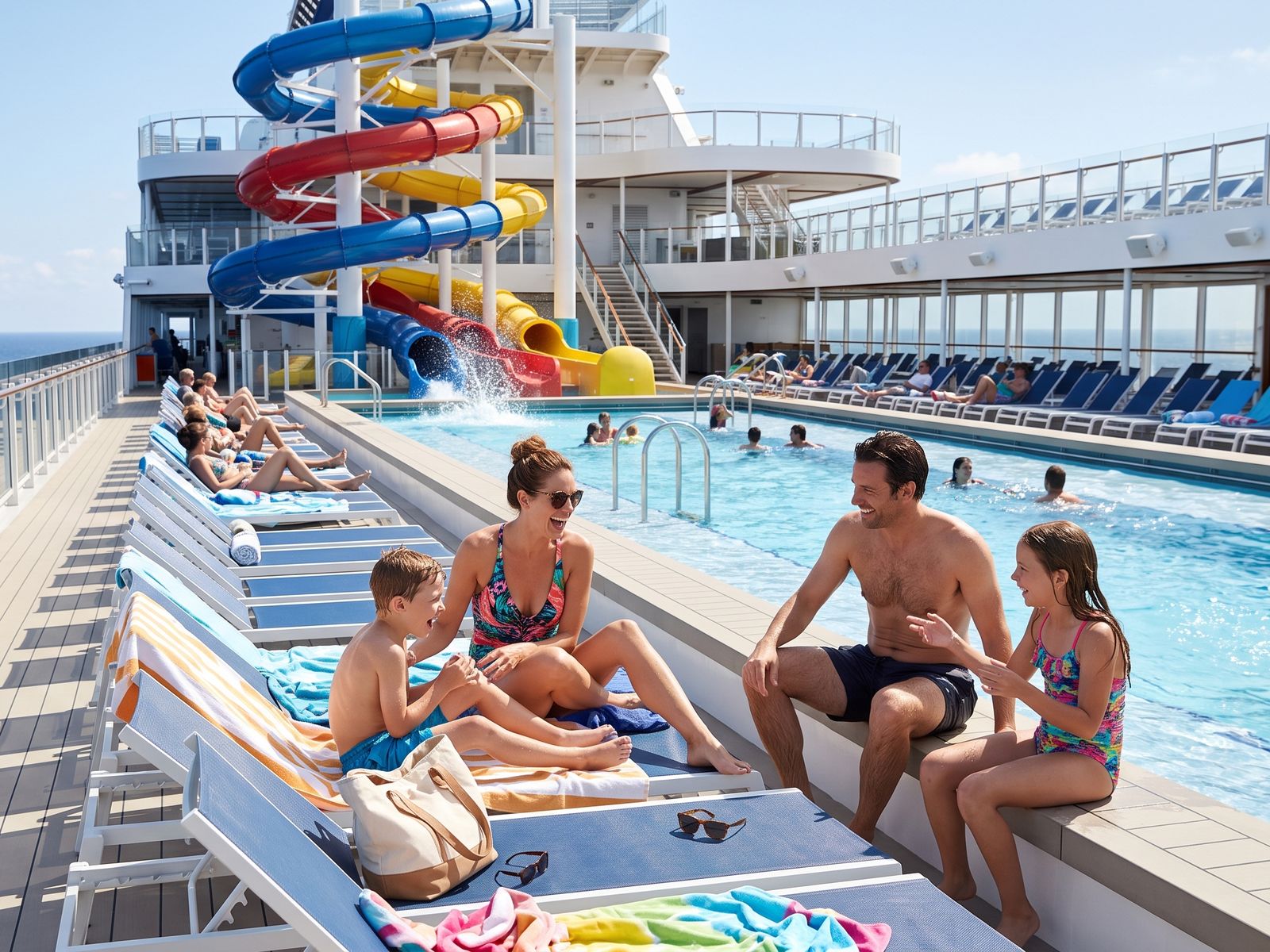Una familia con dos niños se ríe junto a la piscina en la cubierta de un crucero, con toboganes de colores detrás