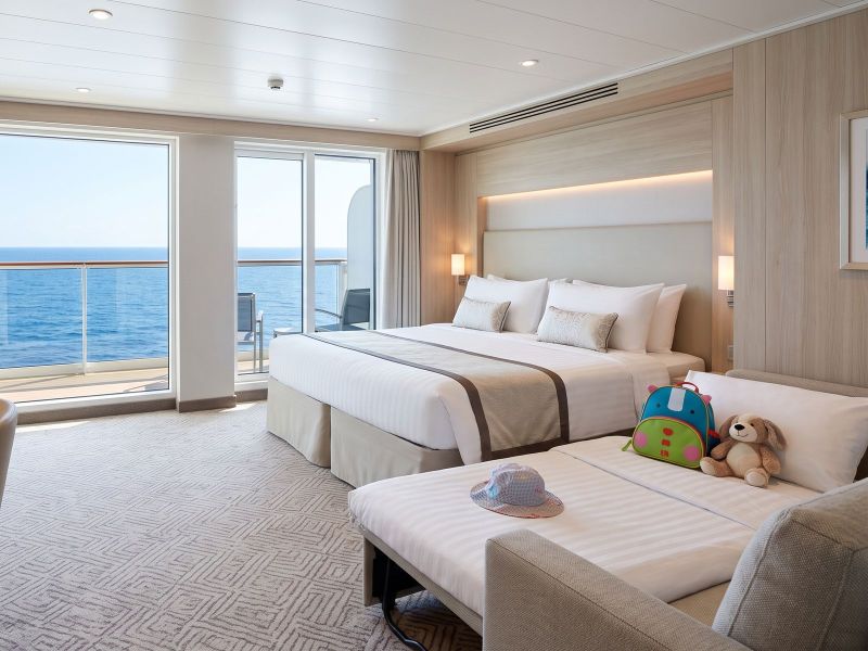 A family cabin with a sea-view balcony: a double bed, a sofa bed made up for a child, a children's backpack and a soft toy